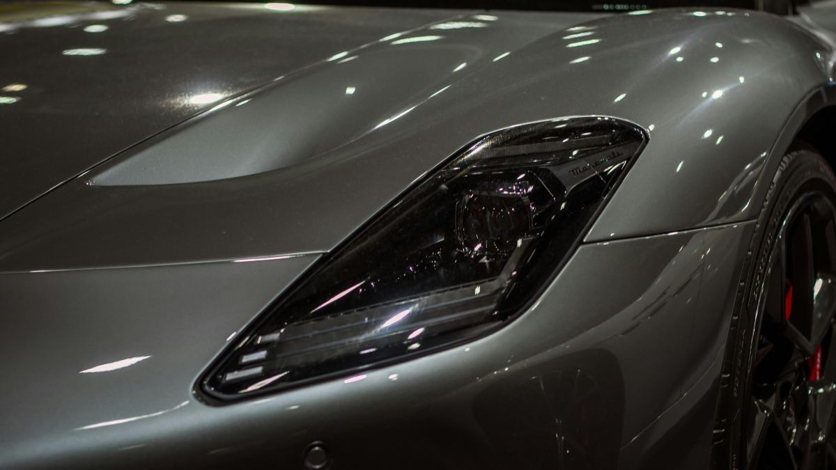 Close-up of a sleek silver sports car's headlight and front fender, glossy paint reflecting overhead lights